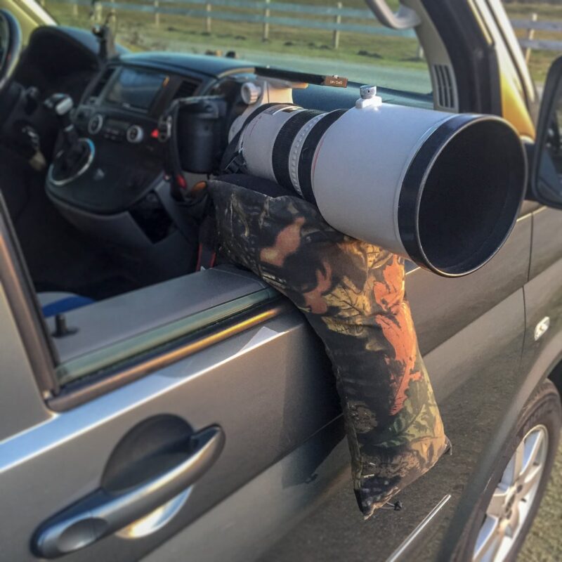 pixelpenguin- Een gecamoufleerde cameralens zittend op een tweepotige zitzak in het raam van een auto.