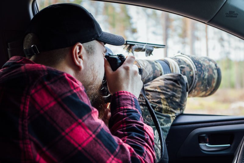 pixelpenguin- Een man in een camouflageshirt maakt een foto uit het raam van een auto met een tweepotige zitzak als ondersteuning.